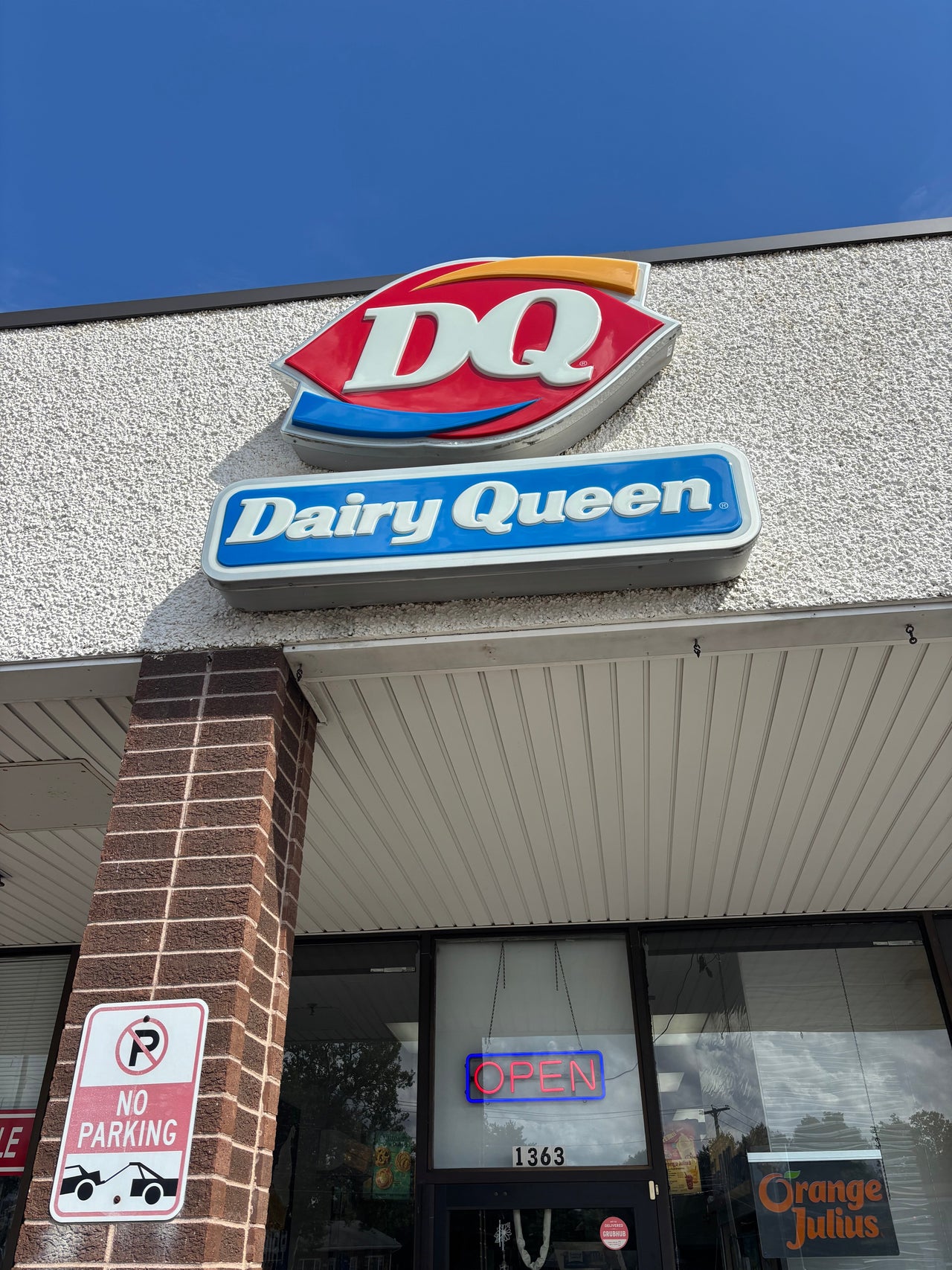 Exterior entrance of a Dairy Queen restaurant with the iconic red-and-blue DQ logo displayed overhead, illustrating a Carpe Diem reflection on simple pleasures, affordable indulgence, and the enduring appeal of the classic Chocolate Dipped Cone.