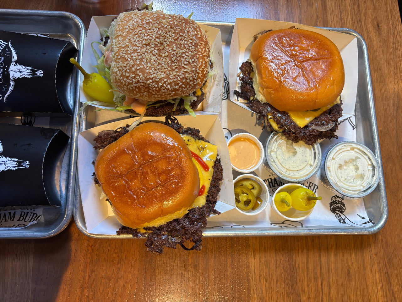 Two Gotham-style smash burgers and a California Dreamin’ burger served with sauces, pickled jalapeños and chili peppers at Gotham Burger Social Club Williamsburg.