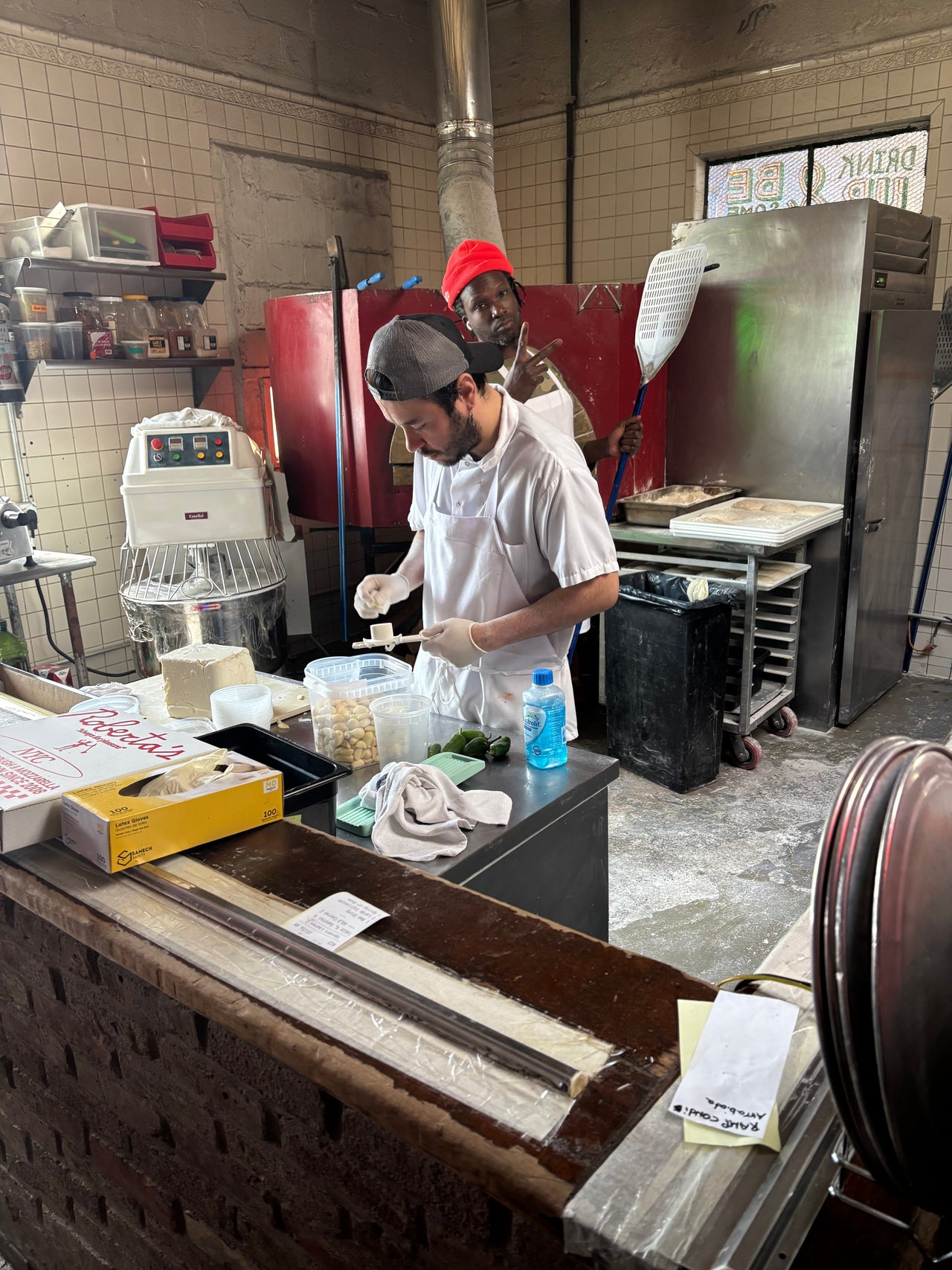 Wood-fired pizzaiolos crafting artisan Neapolitan pizzas and other delights inside Roberta’s bustling open kitchen in Brooklyn, surrounded by flames, fresh dough, and the energetic atmosphere of a busy New York pizza outing.